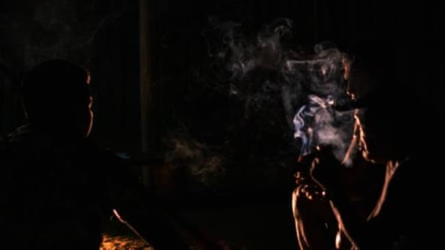 Three men sit in the dark around a fire. One holds a smoking cigar